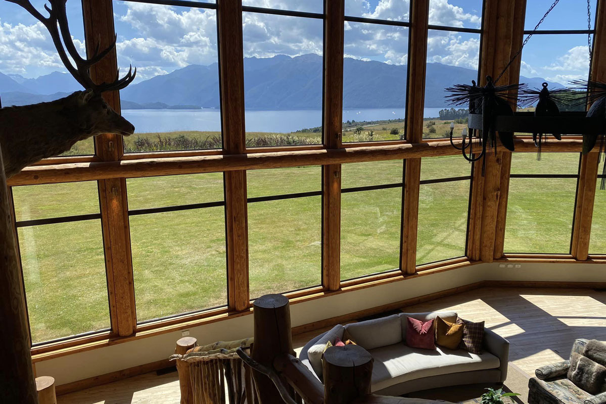 Fiordland Lodge lounge view showing the Great Room interior with comfortable sofas and the full 180-degree panorama of Lake Te Anau and Fiordland National Park mountains visible through the floor-to-ceiling windows