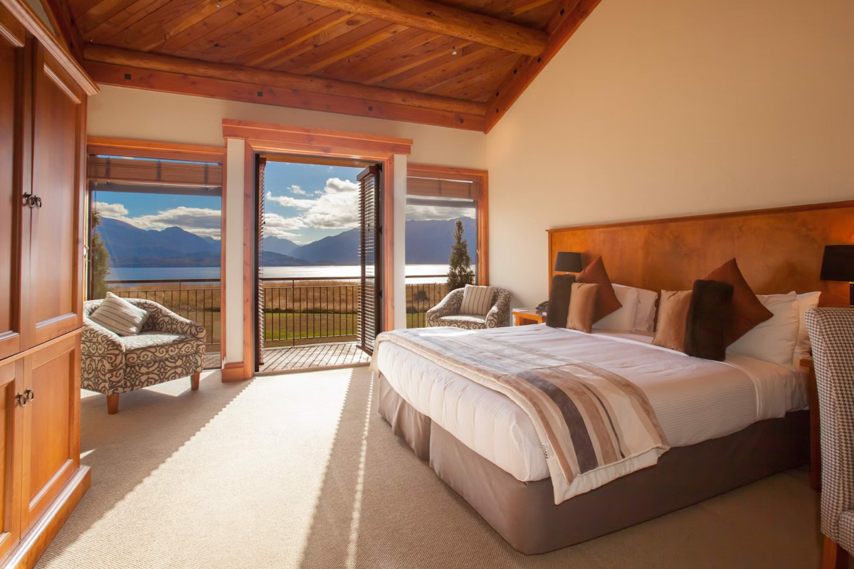 Interior of a Deluxe Lodge Room at Fiordland Lodge showing the king-sized bed with quality linen warm timber walls and large windows overlooking Lake Te Anau and the Fiordland National Park mountain ranges