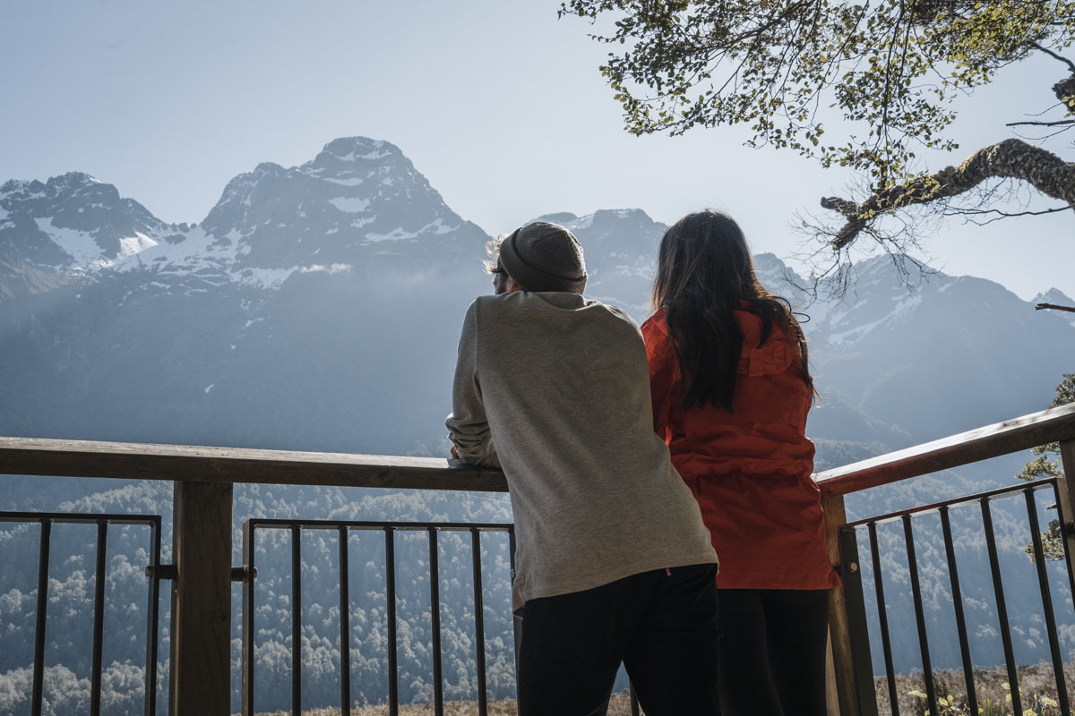 Fiordland Lodge balcony in winter showing a couple on the upper lodge balcony overlooking the snow-dusted Fiordland National Park landscape and the still winter waters of Lake Te Anau below