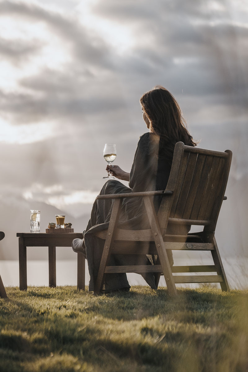 Wine on the terrace at Fiordland Lodge in winter showing a glass of red wine held against a backdrop of Lake Te Anau and the snow-covered Murchison Mountains in fading winter light